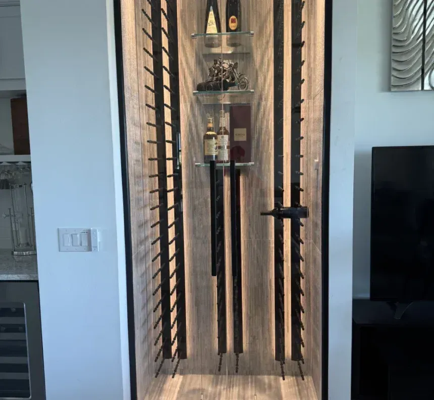 A modern wine display built into a wall, featuring vertical metal bottle racks flanking glass shelves with backlit wood.