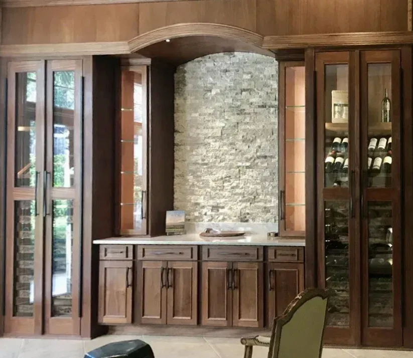 A wet bar featuring wooden cabinetry, a stone accent wall, and glass-front wine storage under an arched canopy.