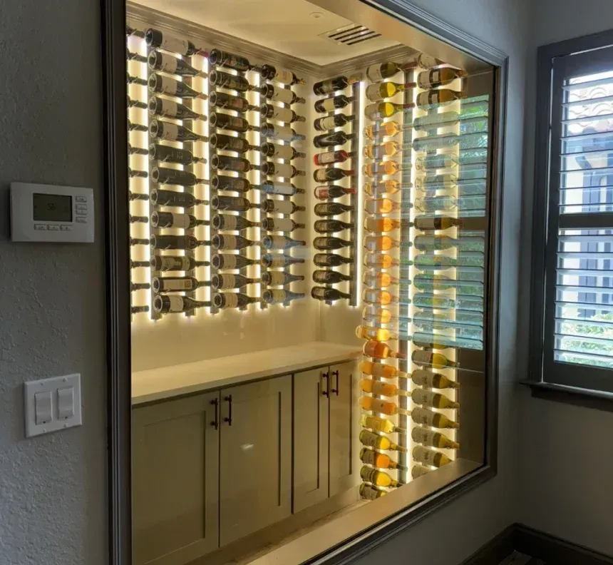 Built-in wine cellar with illuminated vertical bottle racks above white cabinets behind a glass wall.