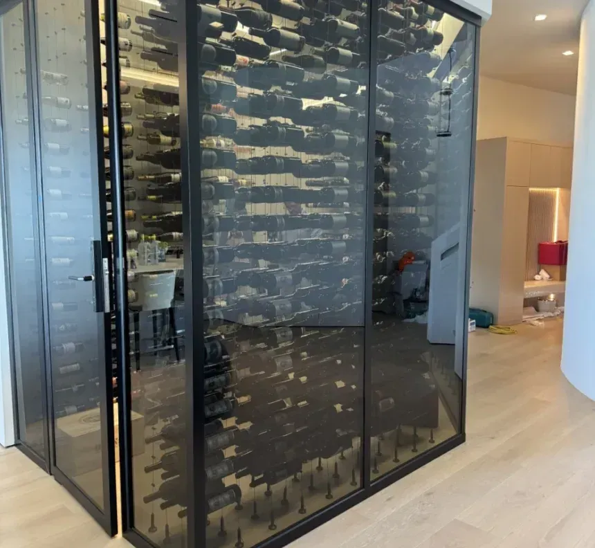 A modern, floor-to-ceiling glass wine cellar with black metal framing, displaying rows of wine bottles in a home interior.