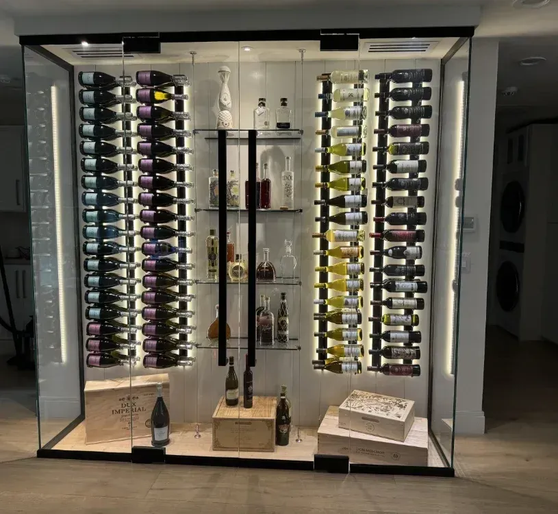 A glass-enclosed, illuminated wine cellar with shelves holding bottles, centered display racks, and wooden storage boxes.