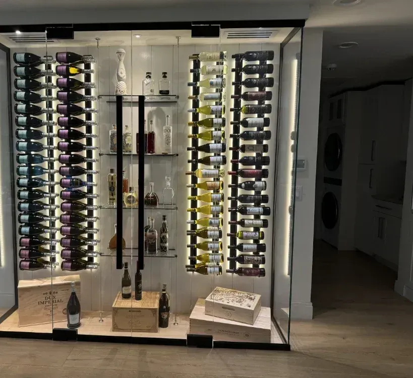 A modern, backlit glass wine cellar featuring vertical bottle racks, glass shelves with liquor bottles, and wooden crates.