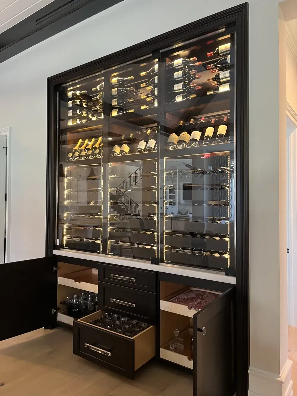 A wall-mounted, black-framed wine display cabinet with interior lighting, glass shelving, and lower storage drawers.