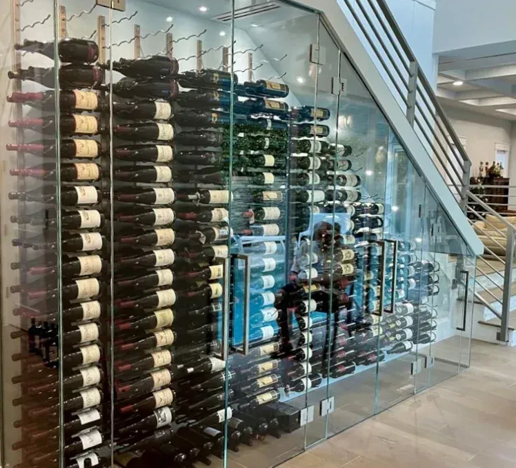 A glass-enclosed wine cellar built under a staircase, filled with rows of bottled wine on horizontal metal racks.