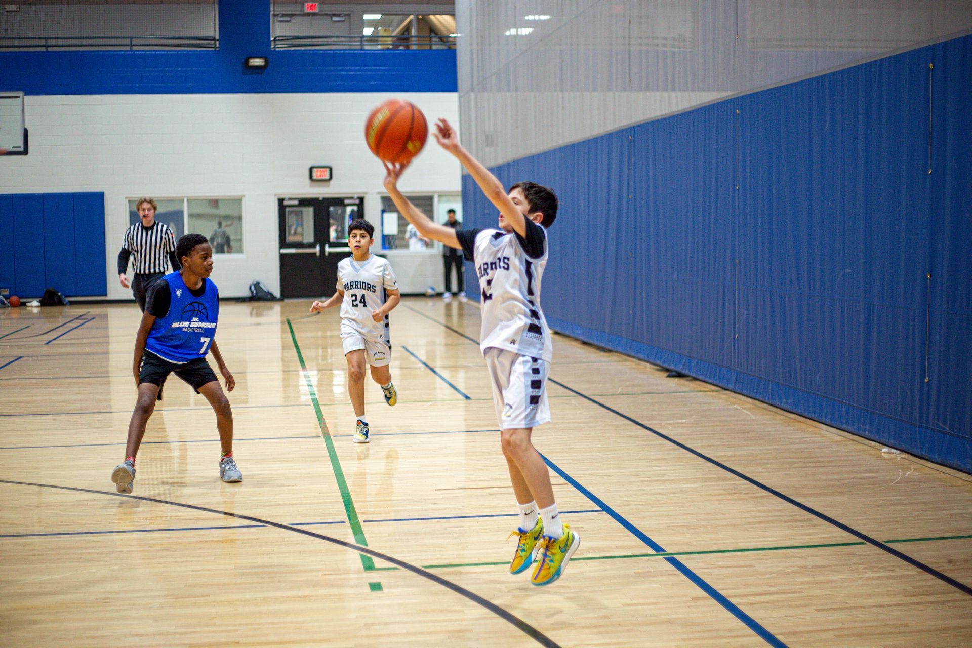 Basketball player in white shooting a jump shot while a defender in blue watches in a gym