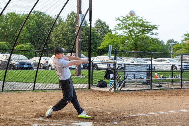 Softball player in white swings a bat on a dirt field near a chain-link fence.