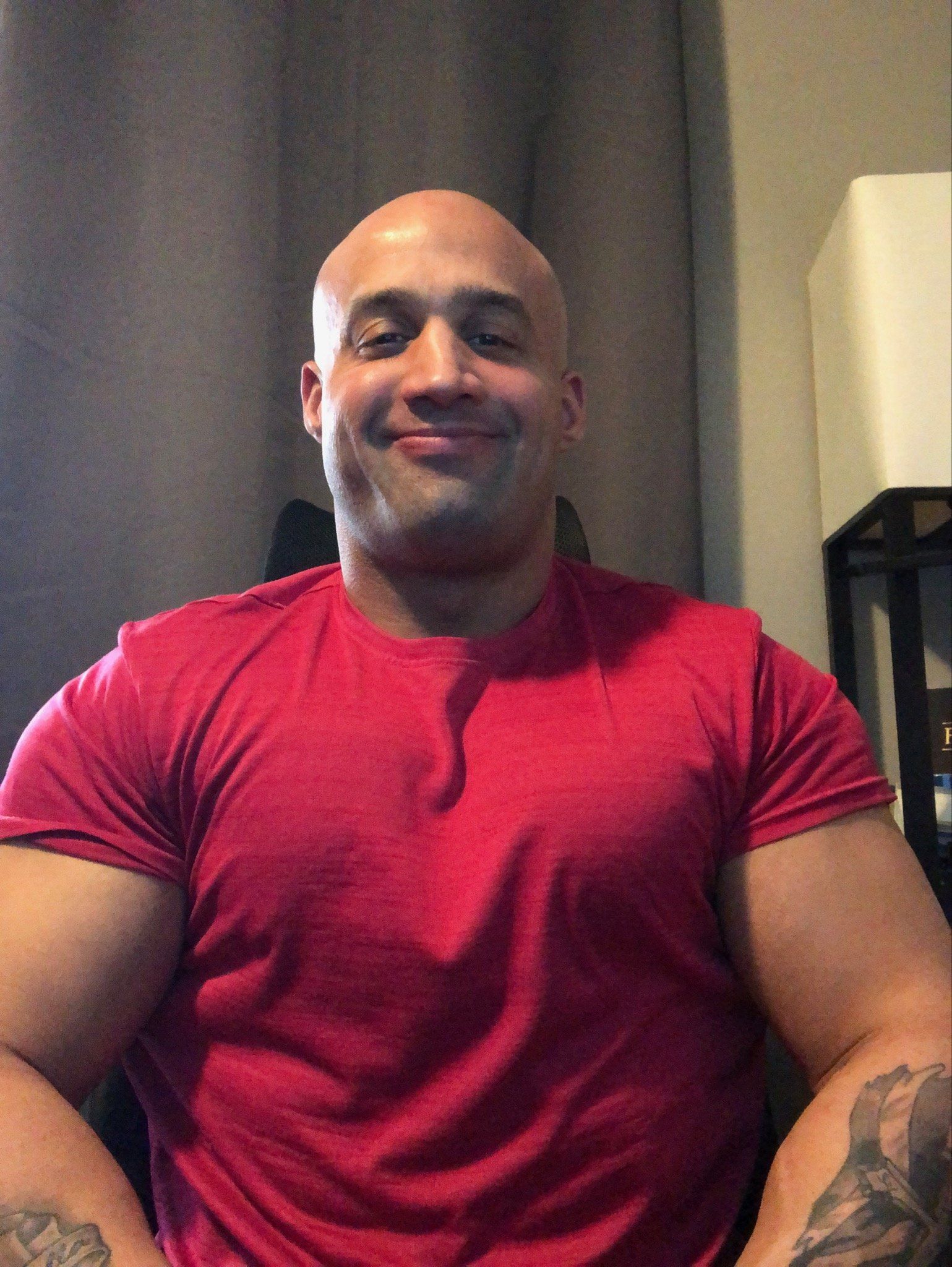 A man with a tattoo on his arm is wearing a red shirt and smiling.