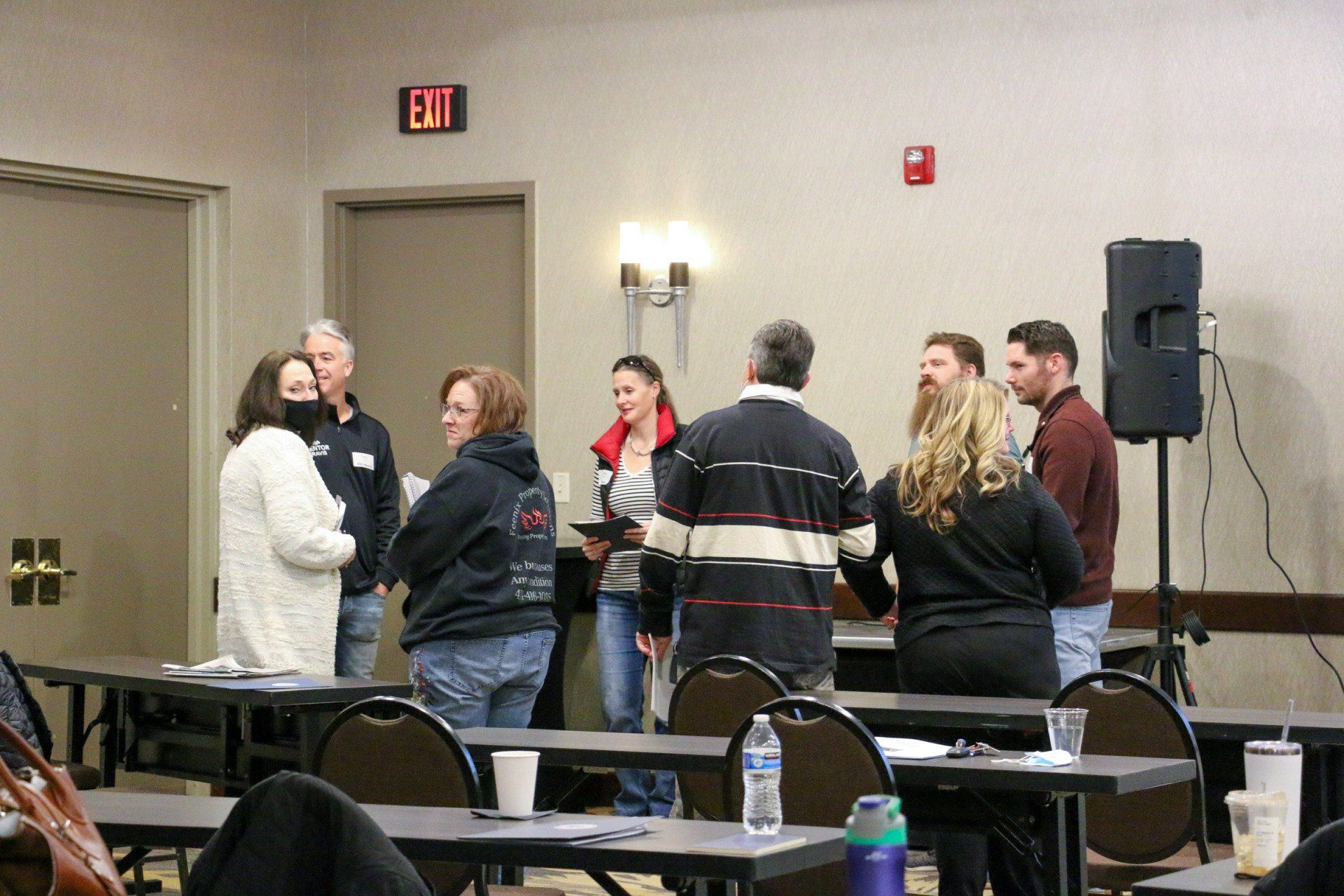 A group of people are standing around tables in a room with an exit sign.