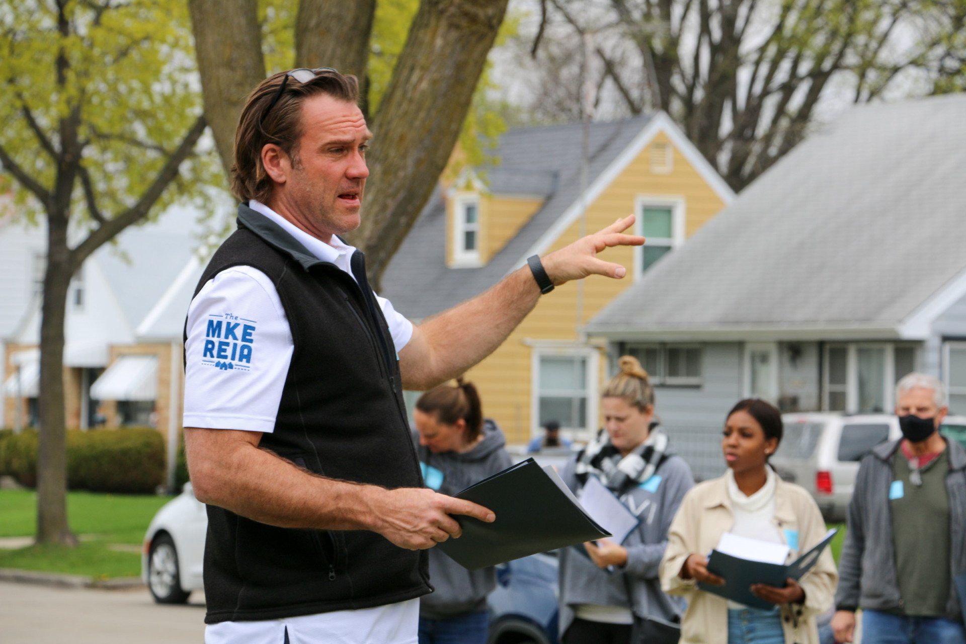 A man is holding a clipboard and pointing at something while standing in front of a group of people.