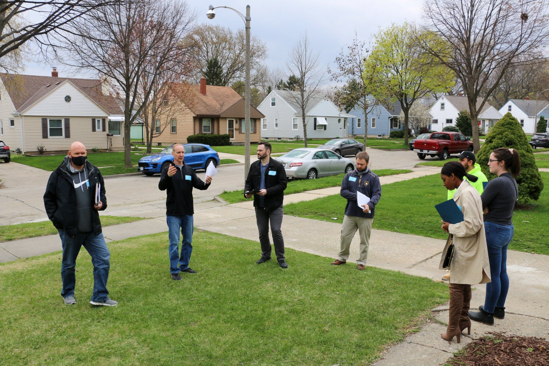 A group of people are standing on a lush green lawn in a residential neighborhood.