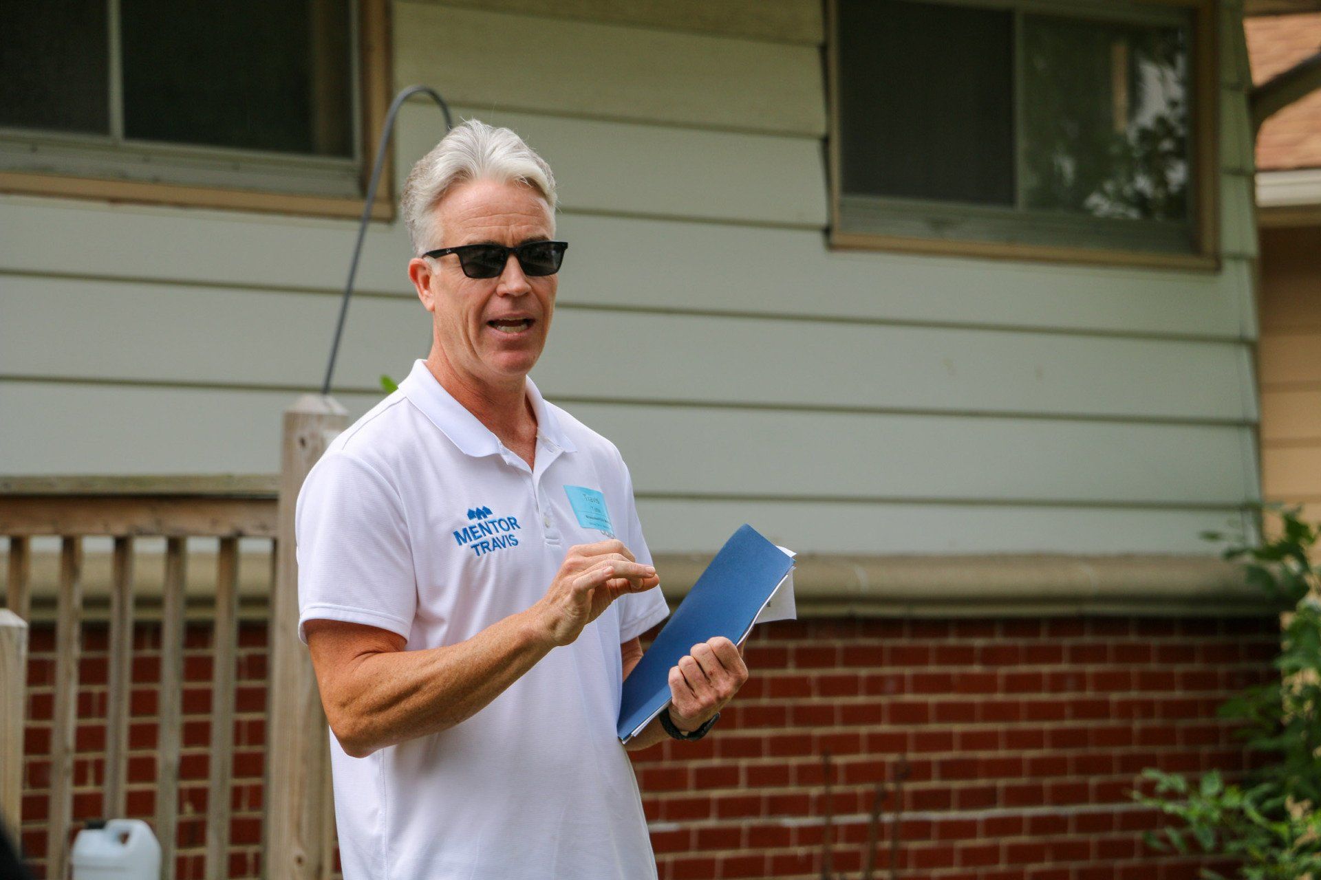 A man wearing sunglasses and a white shirt is standing in front of a brick house holding a clipboard.