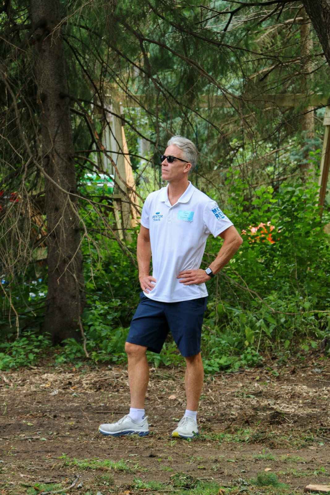 A man in a white shirt and blue shorts is standing in the woods.