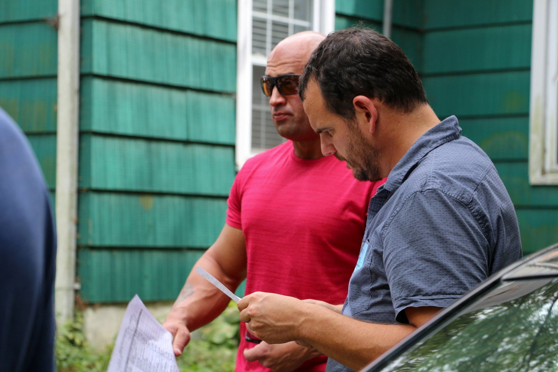 Two men are standing in front of a green house looking at a piece of paper.