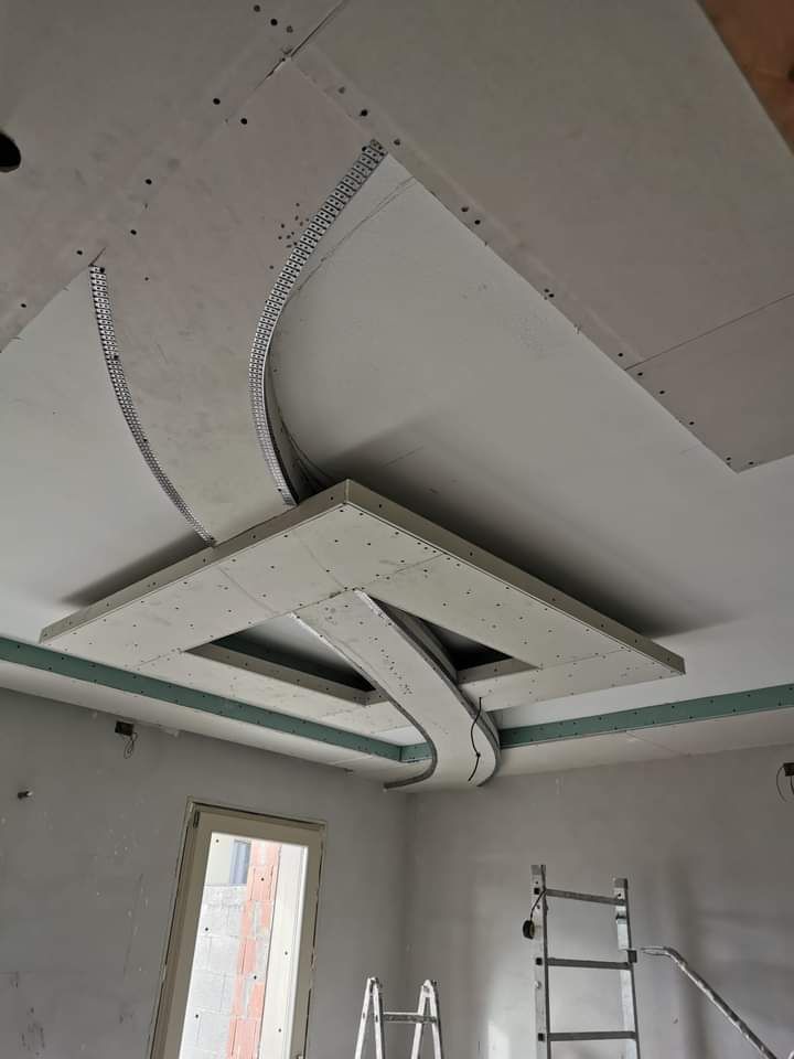 Soffitto in costruzione con pannelli in cartongesso, aperture curve e una scala sottostante.
