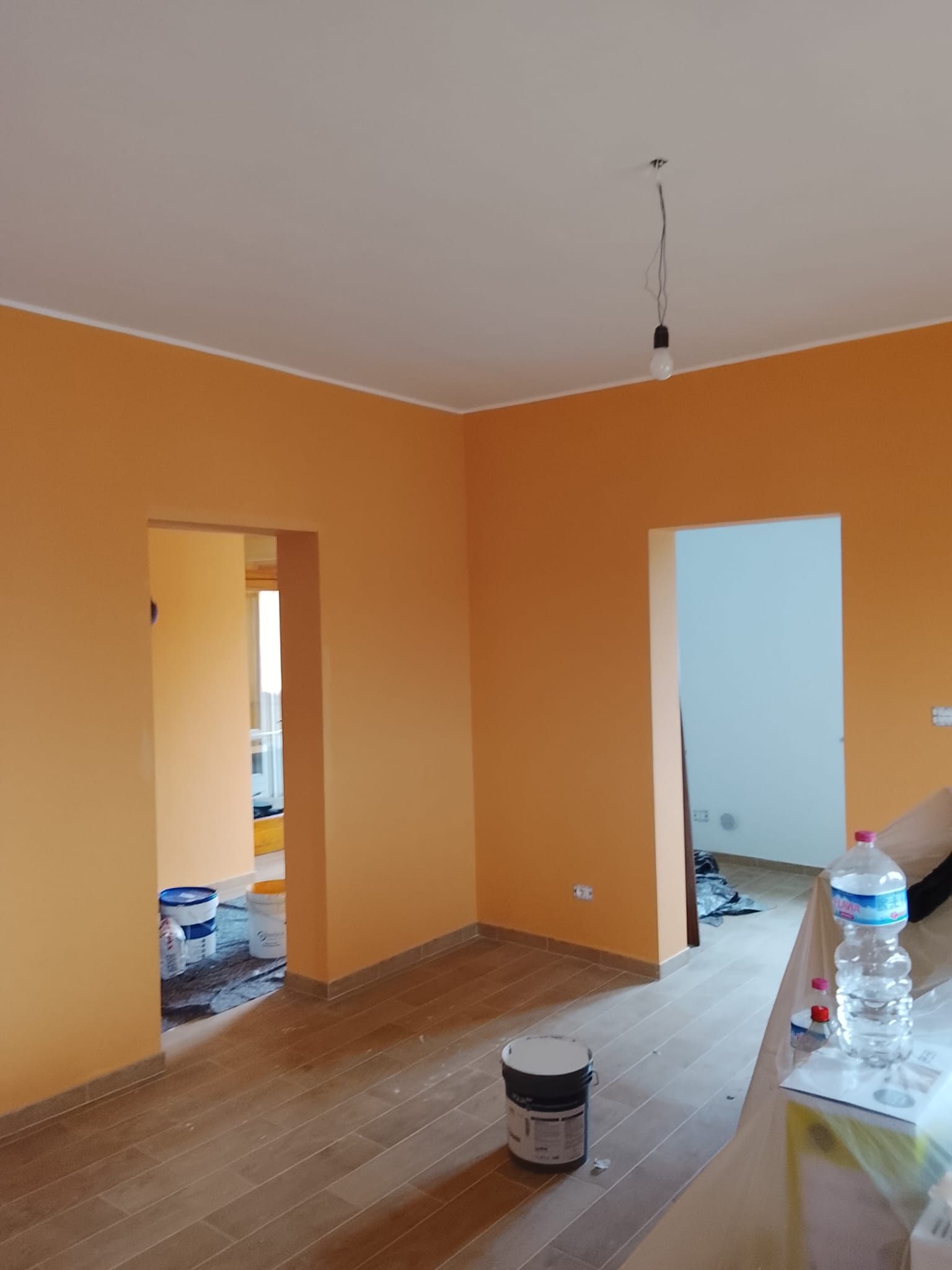 Stanza vuota dipinta di arancione, in fase di ristrutturazione, con pavimento grezzo, due porte e un secchio di vernice sul pavimento.