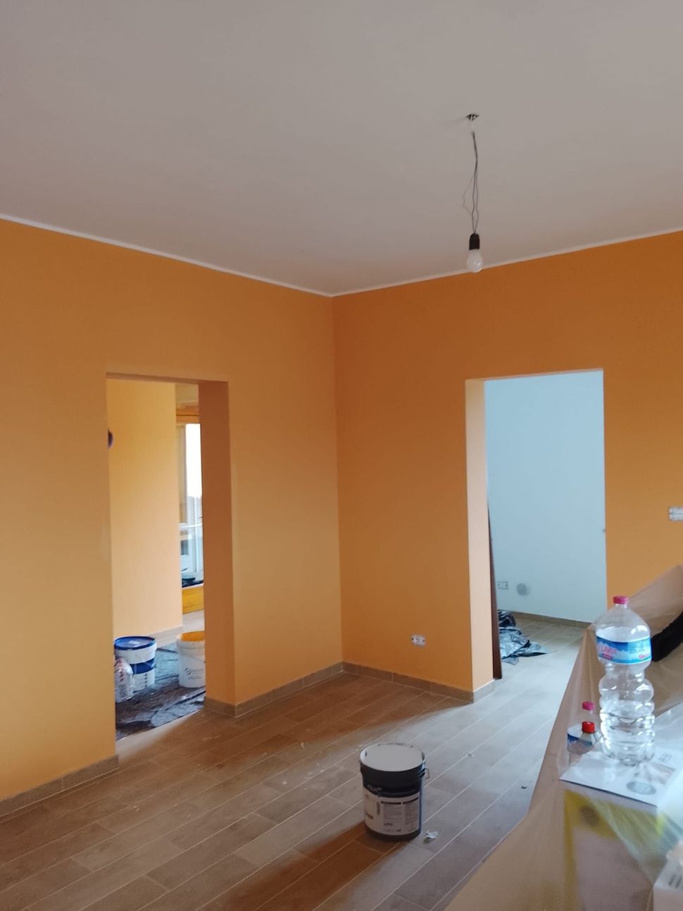 Stanza dipinta di arancione in fase di ristrutturazione, con porte, una lampadina appesa e materiali per la pittura sul pavimento.