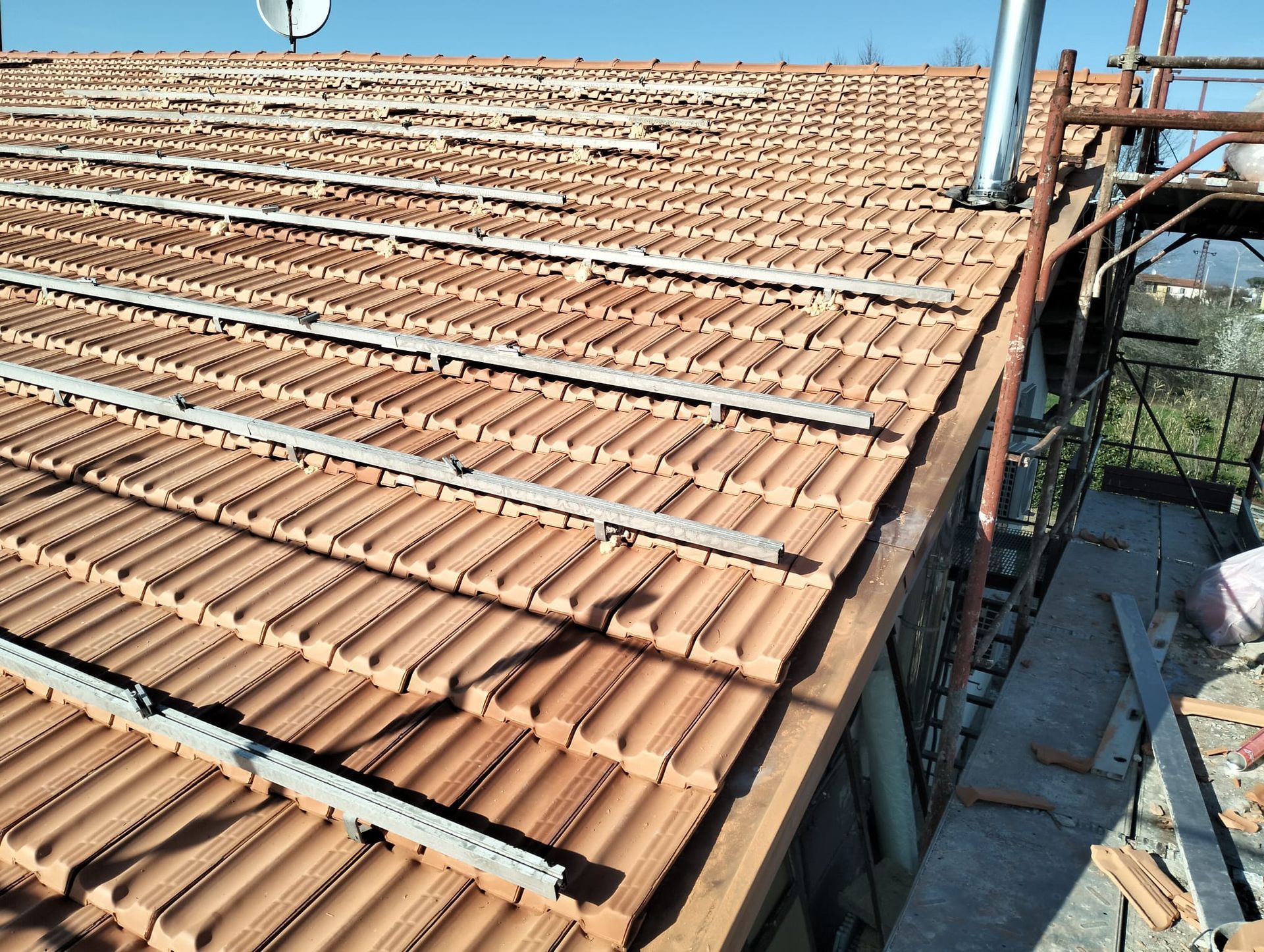Tetto in costruzione con tegole bruno-rossastre, con impalcature e cielo azzurro a fare da sfondo.