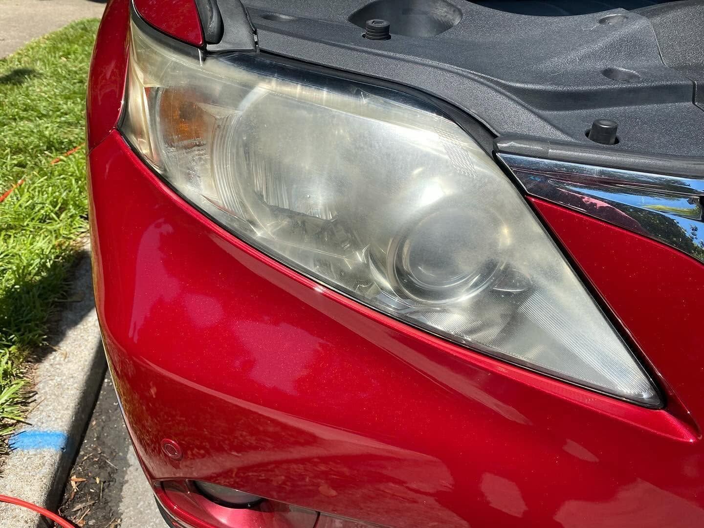 Close-up of a red car’s front headlight and bumper with hood open outdoors
