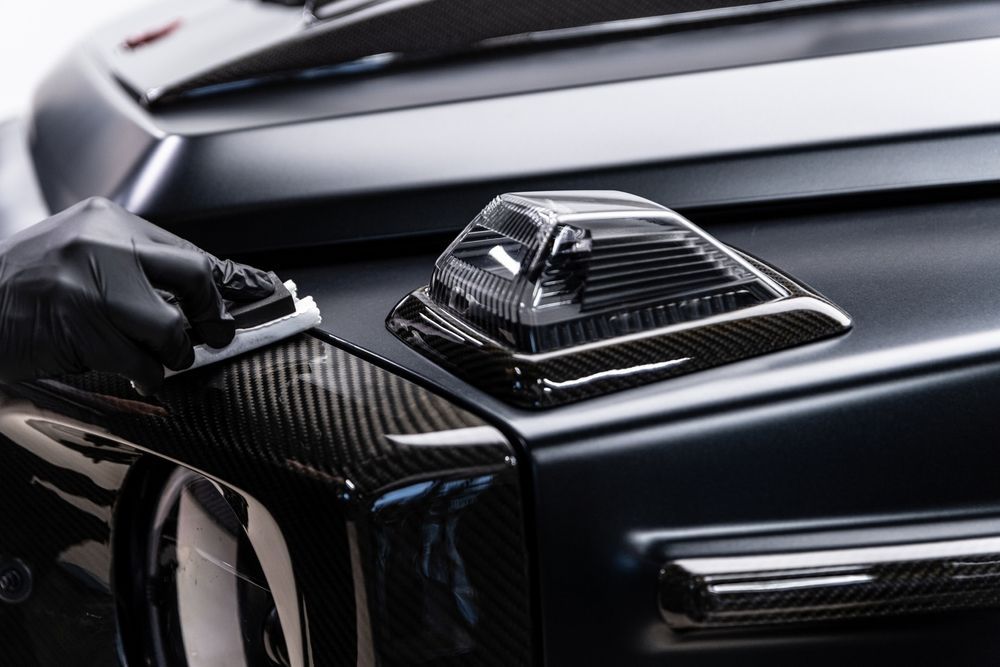 Gloved hand polishing a black carbon-fiber car hood with a glossy air vent detail