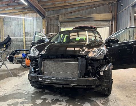 Black car with front damage and missing bumper in a repair garage, another car parked beside it