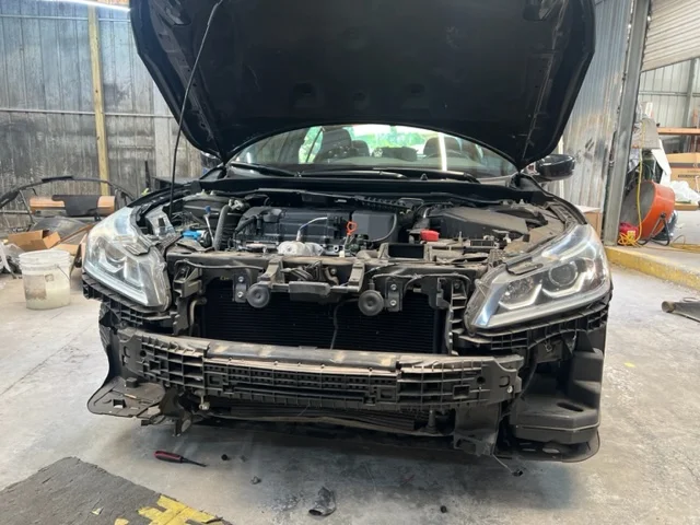 Black car with open hood and front bumper removed in a repair shop.
