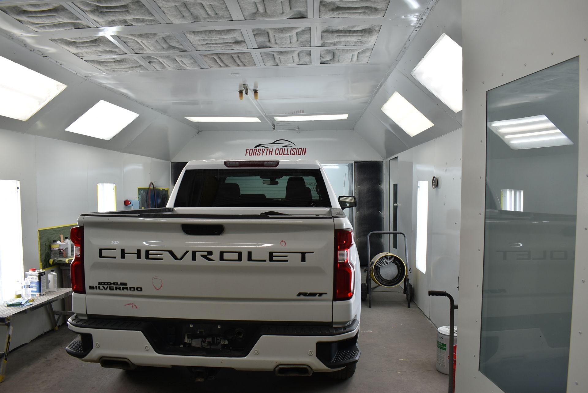 White Chevrolet pickup in an auto body paint booth with bright ceiling lights and open doors
