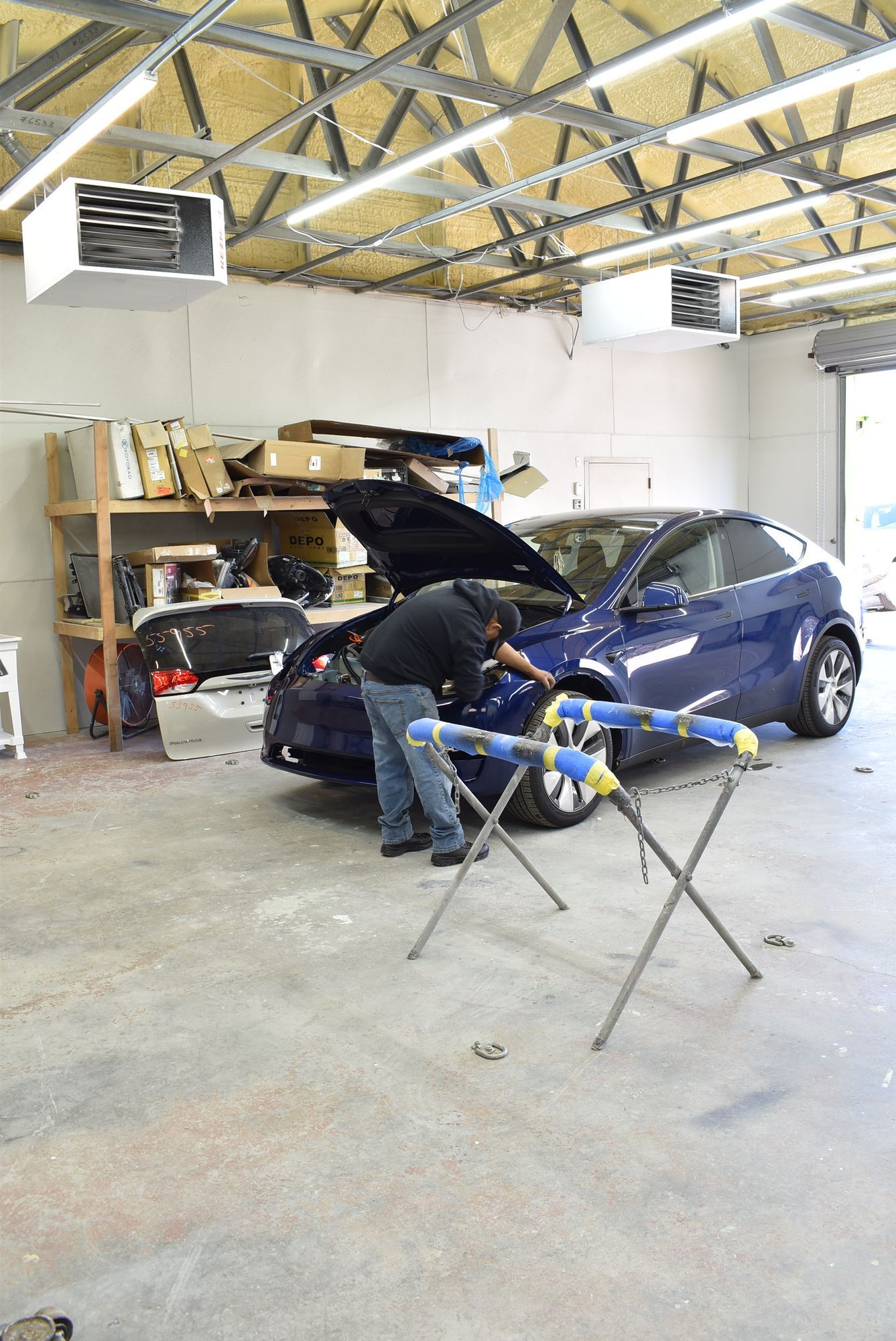 Mechanic working on a blue car in a garage with its hood open, shelves and tools in the background