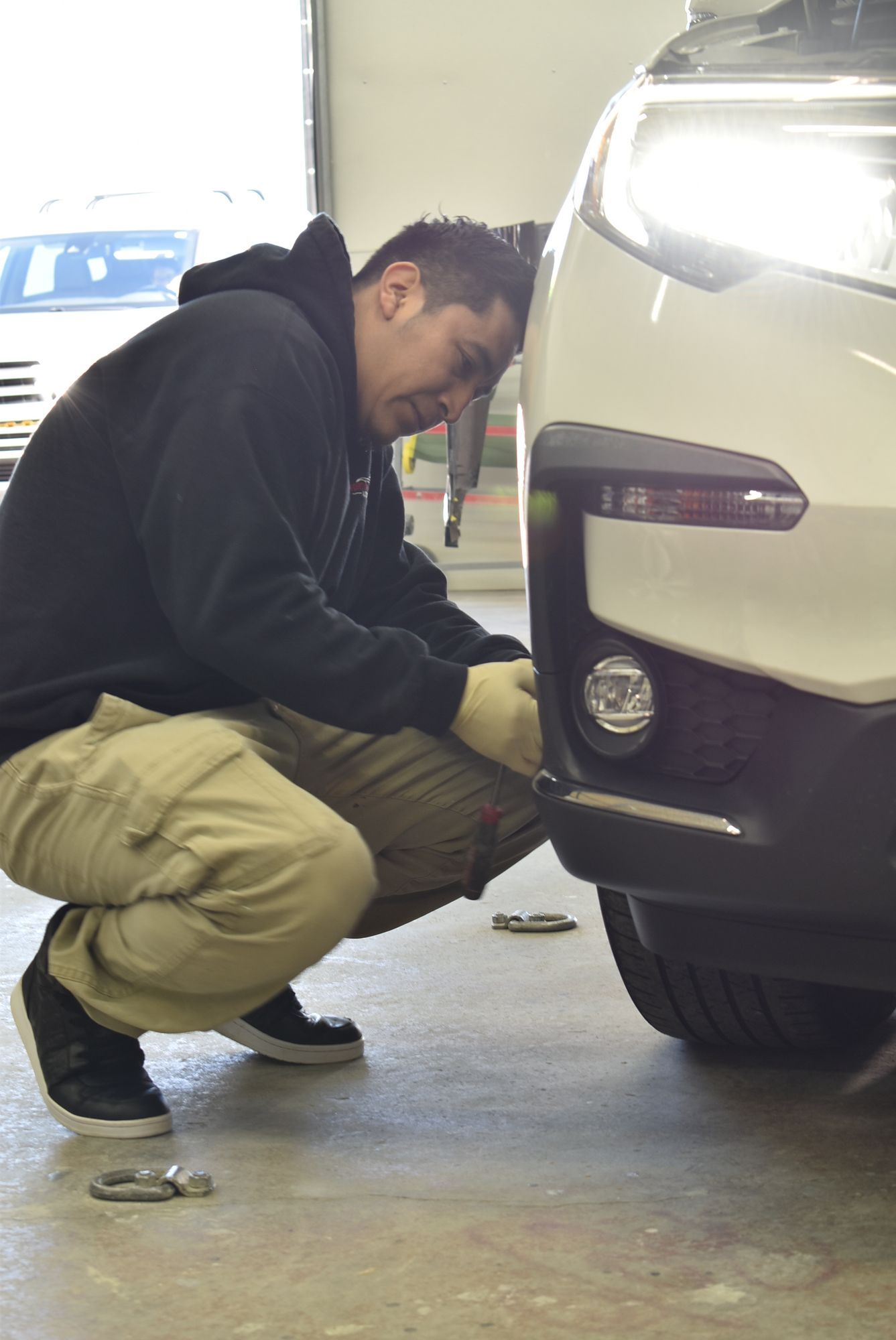Mechanic crouching beside a white car, inspecting the front bumper in a garage