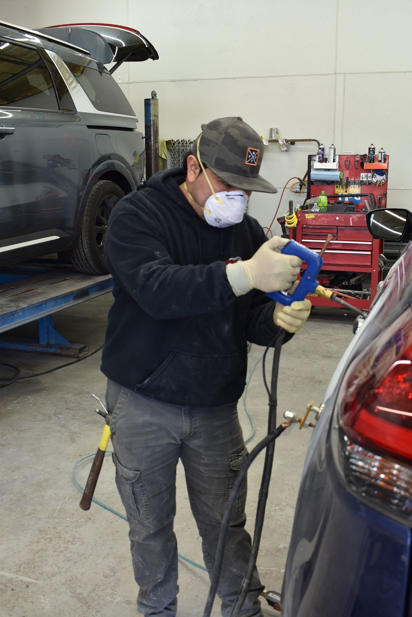 Mechanic using a blue handheld tool beside a dark SUV in an auto repair shop