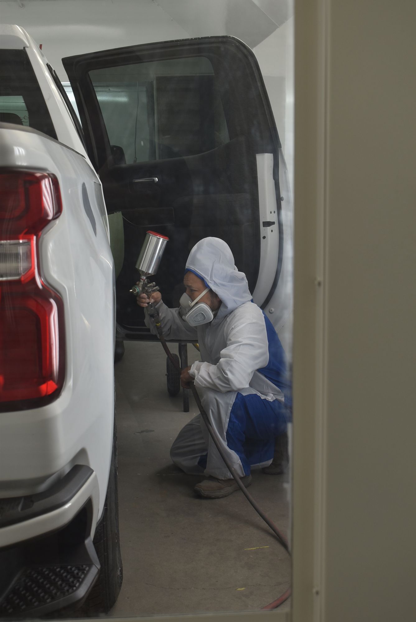 Person kneeling in a garage sprays paint on a car door beside a white SUV