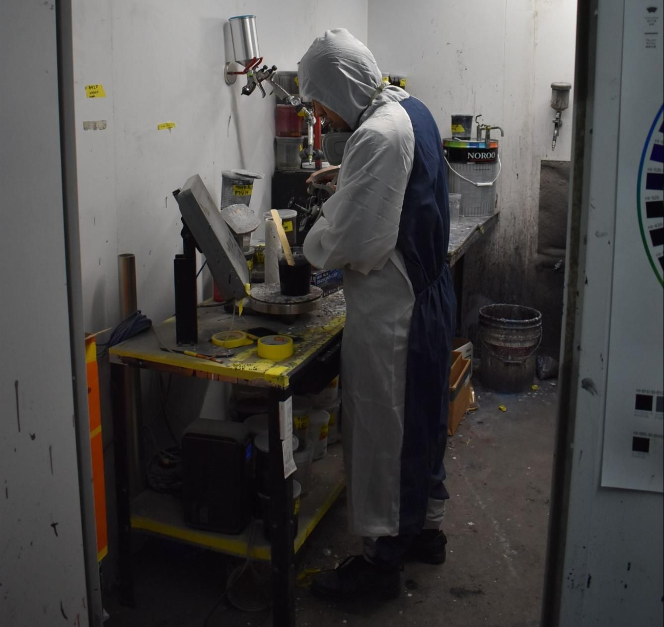A person in a protective suit works with equipment at a metal workbench inside a small, utilitarian workshop.