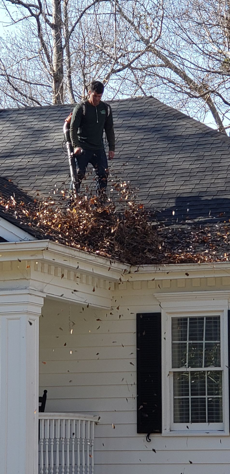A man is standing on top of a roof raking leaves.