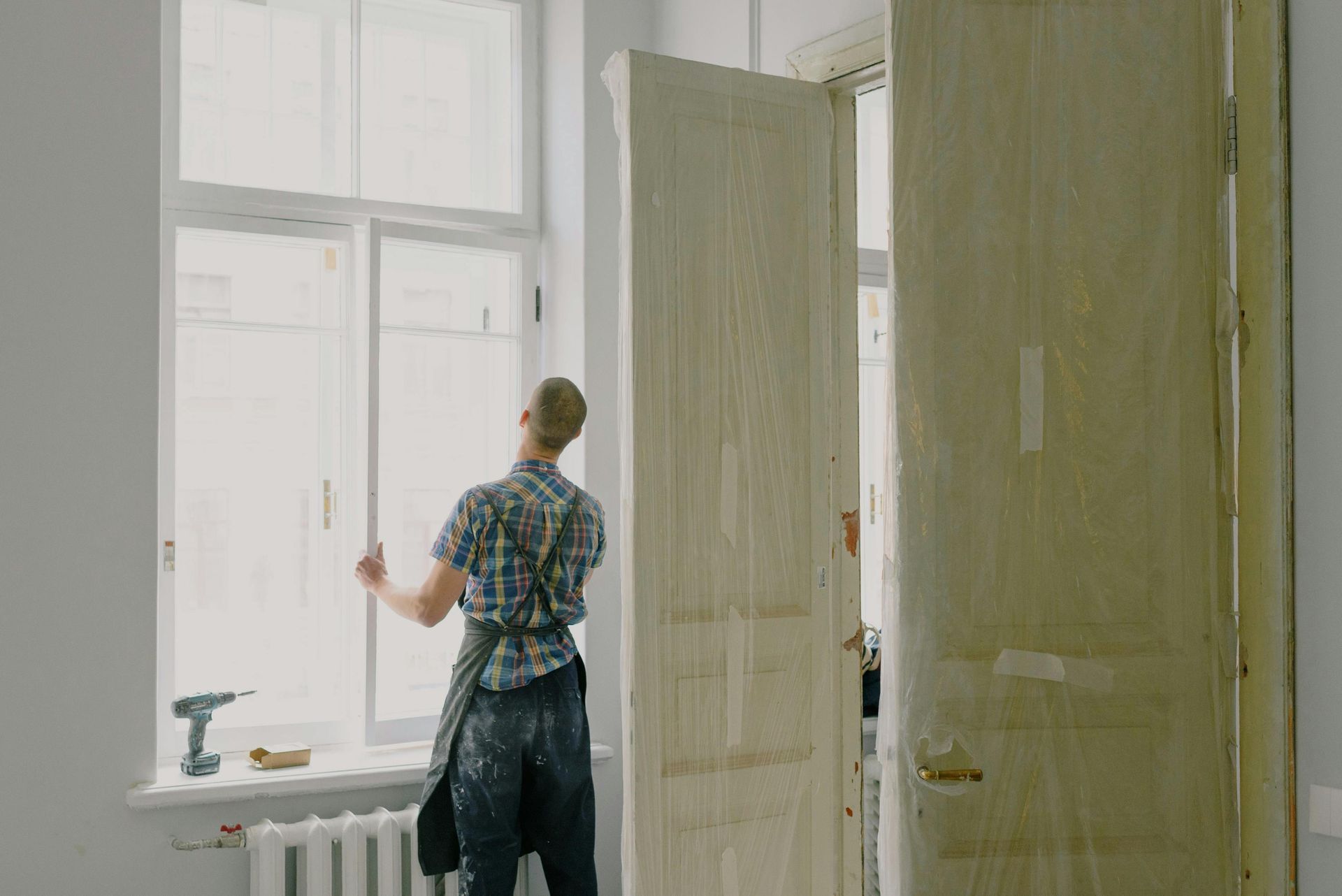 A person wearing an apron works on a window frame in a room next to a large, light-colored wooden door.