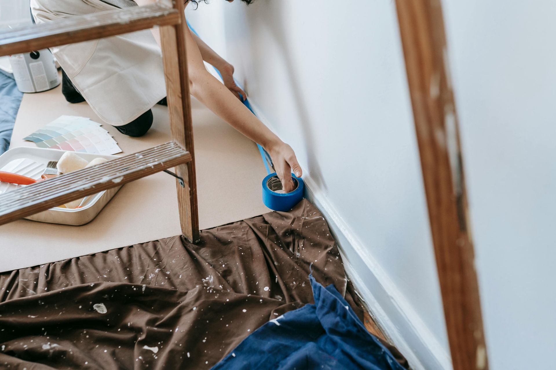 A person kneeling on a floor cloth applies blue painter's tape to the edge of a white wall for a painting project.