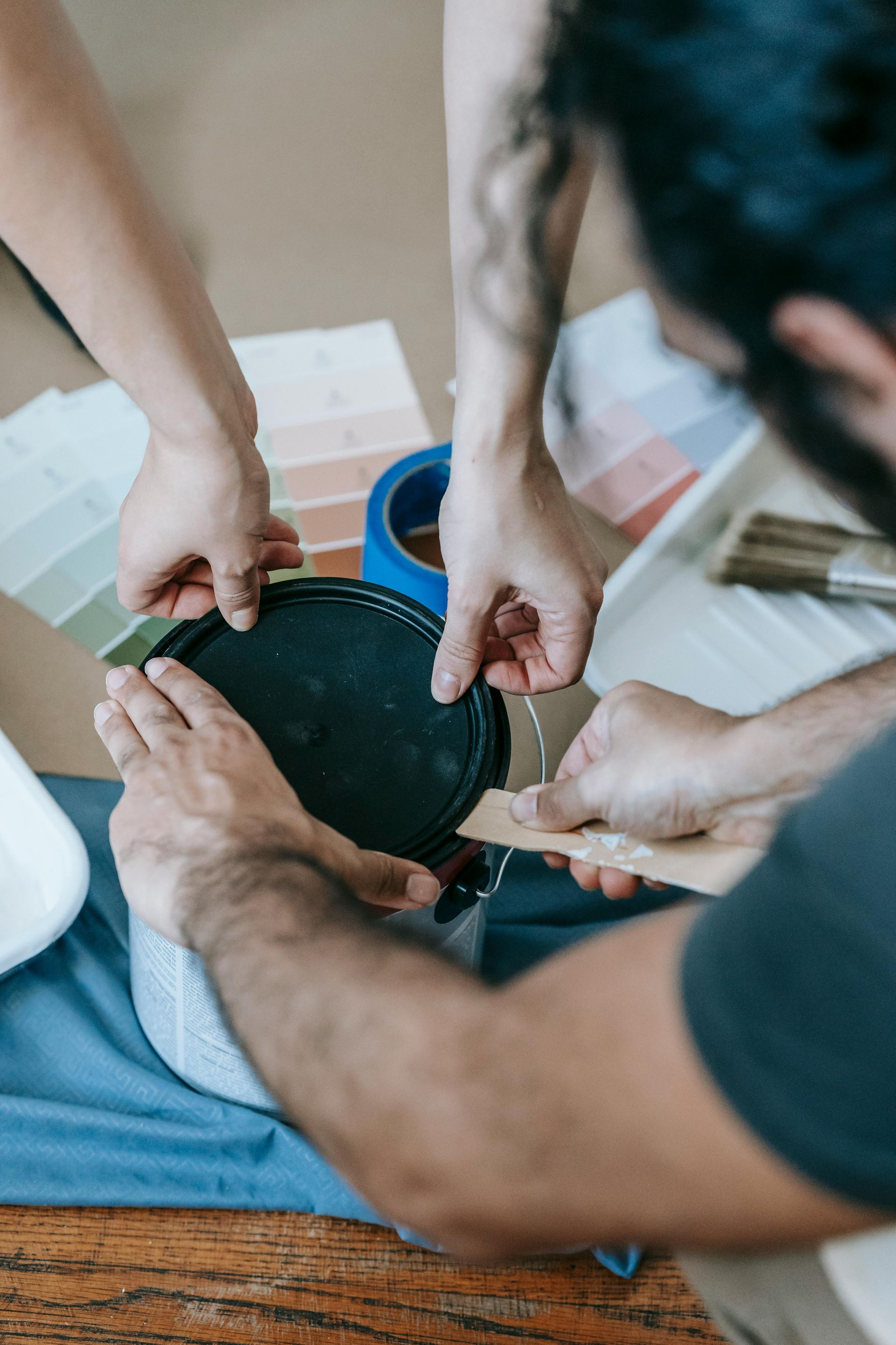 Multiple pairs of hands work together to pry open a paint can on a surface with paint swatches and a paint tray.