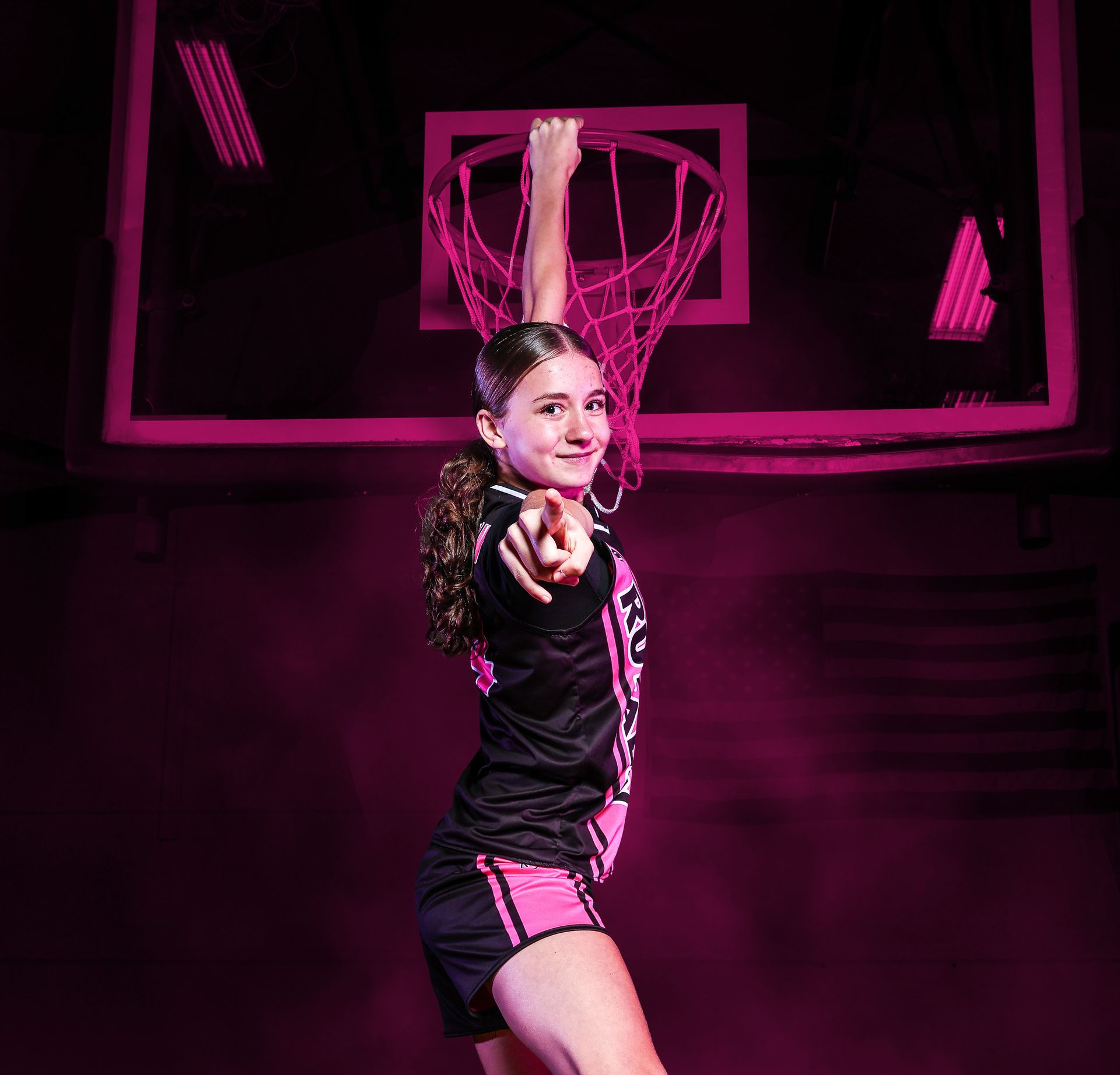 A basketball player jumps to reach a hoop, pointing toward the viewer, bathed in vibrant pink lighting.