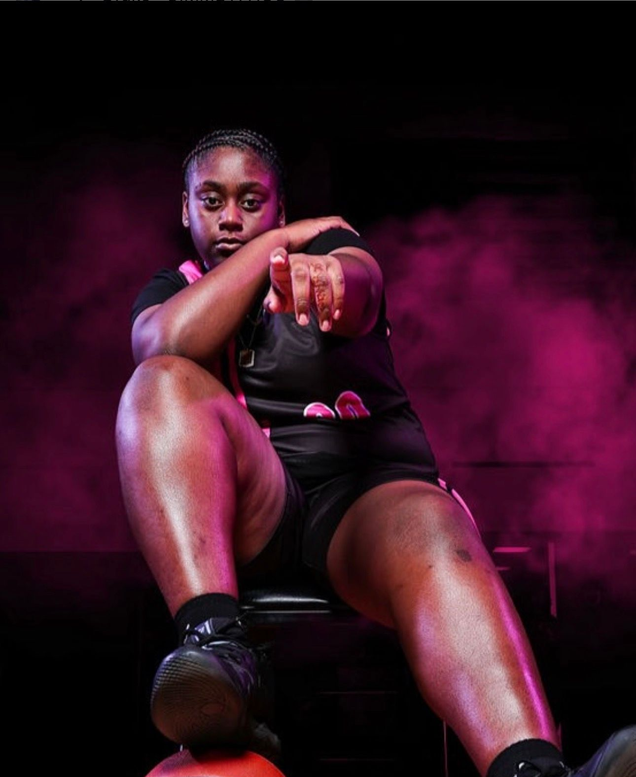 A basketball player in black jersey sits, pointing forward with one foot resting on a basketball against a purple backdrop.