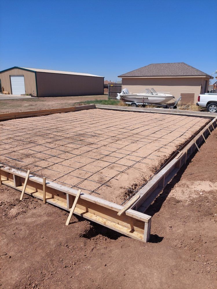 Concrete foundation slab under construction on a dirt lot, with forms, rebar, and houses in the background