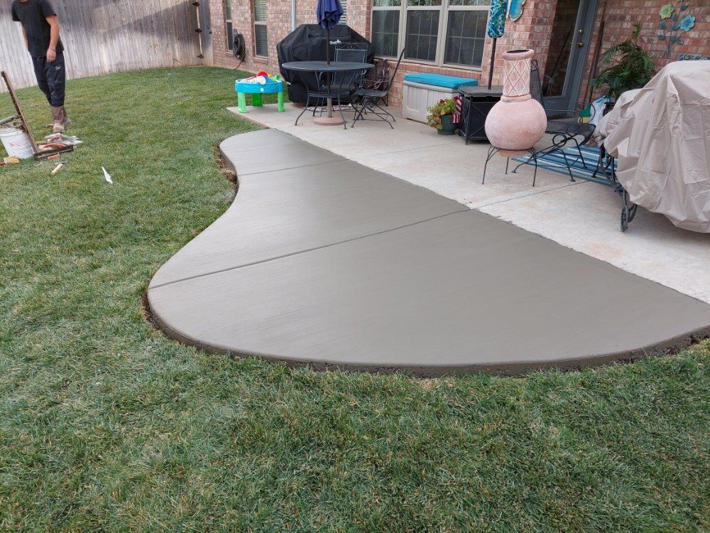 Curved beige concrete patio extension in a backyard with grass, grill, and covered furniture