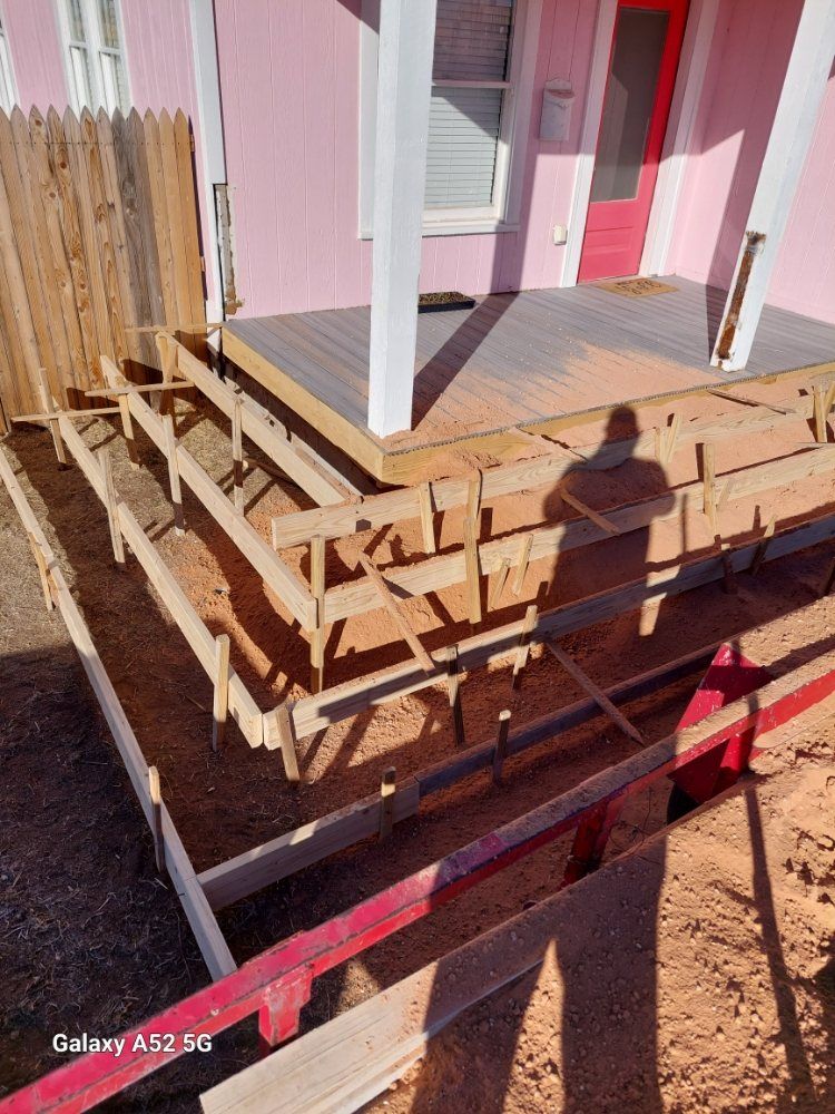 Front porch under construction with stacked lumber, steps, pink siding, and a red door in sunlight