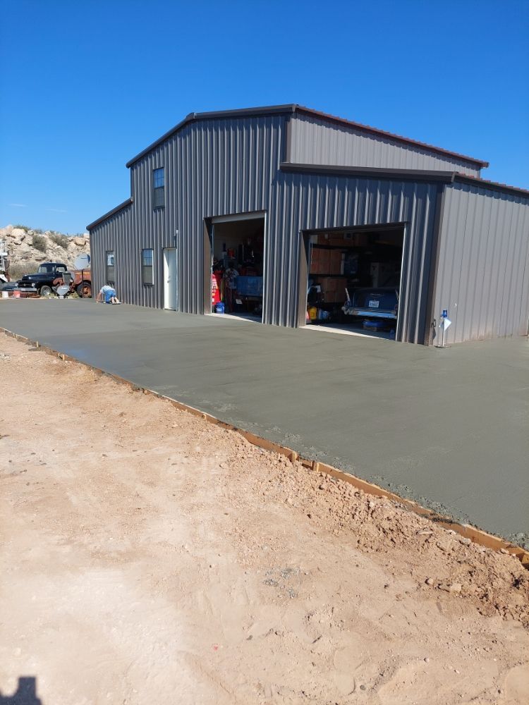 Gray metal garage workshop with open bay doors and paved driveway under a clear blue sky