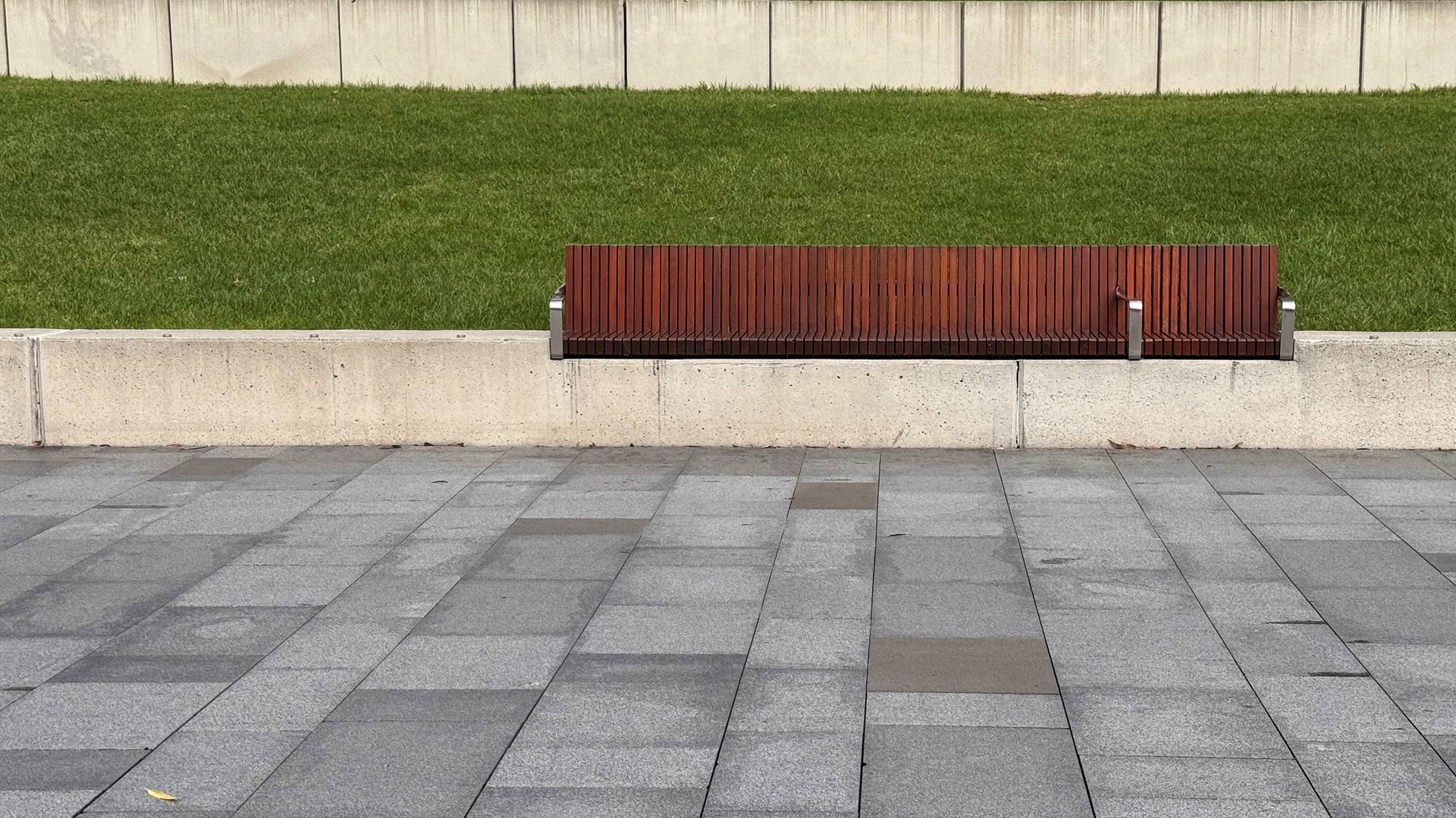 Wooden bench against a concrete wall beside a green hedge on a gray paved plaza.