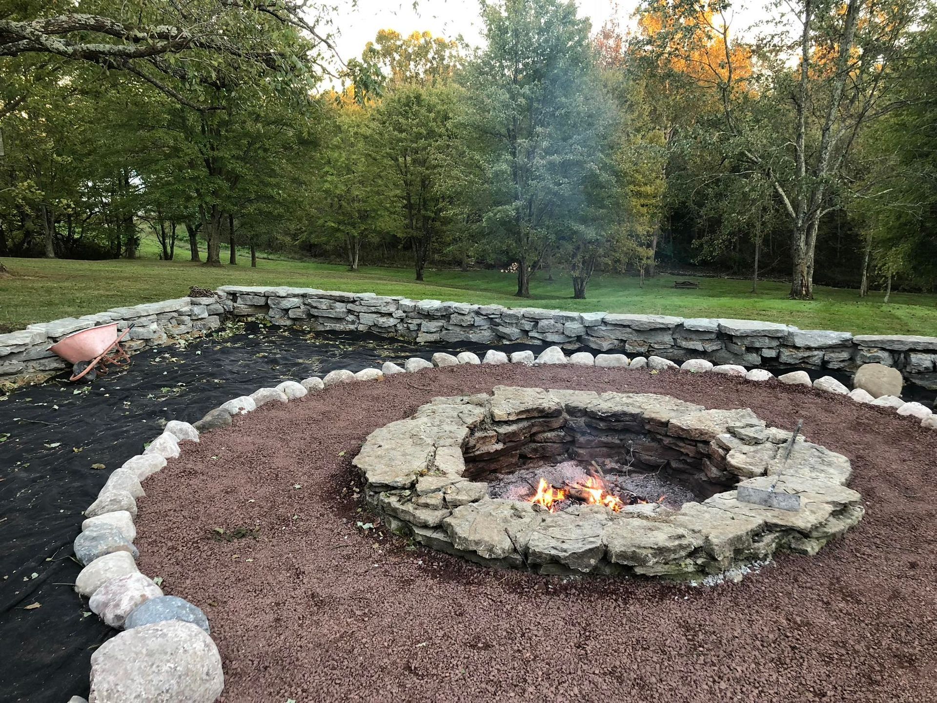 Stone fire pit with burning flames, surrounded by stones and mulch. A stone wall is behind it, and trees are in the background.