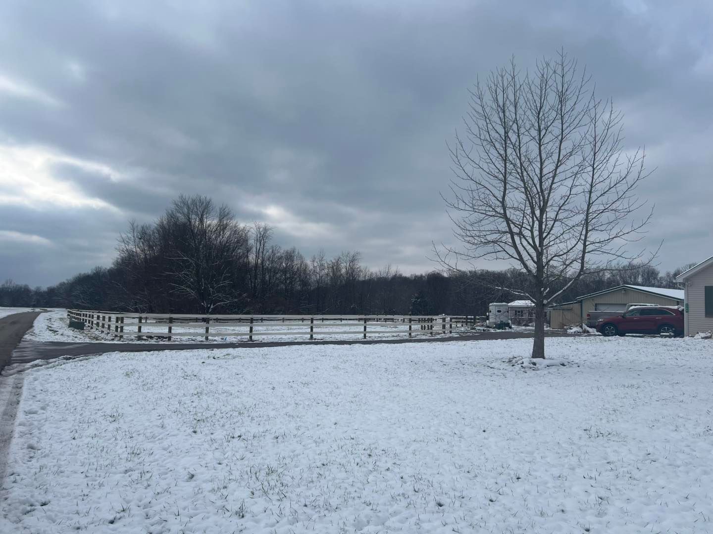 Snowy landscape with a bare tree, fence, and a building on a cloudy day.