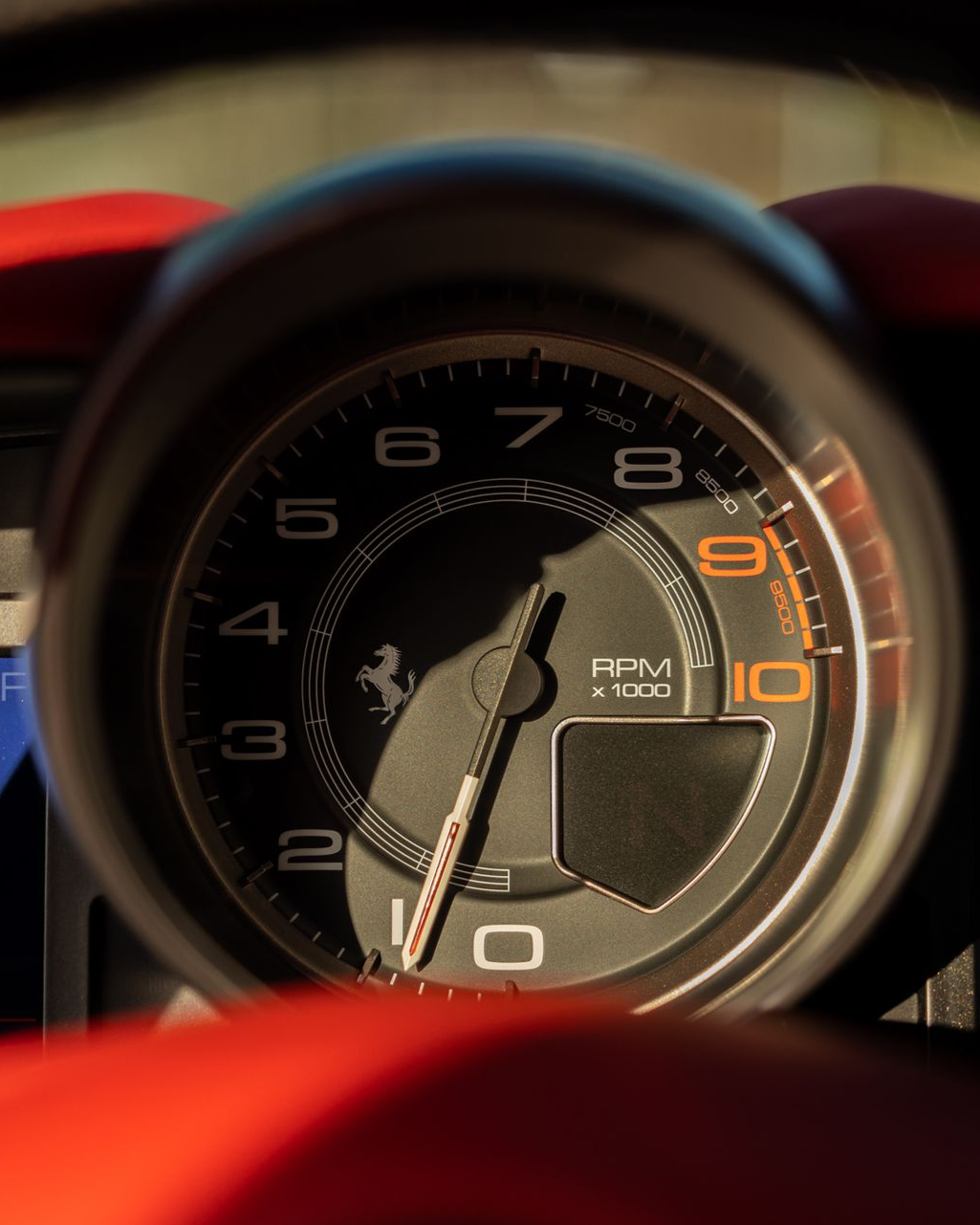 Close-up of a Ferrari dashboard tachometer with red trim, needle near 9,000 RPM.