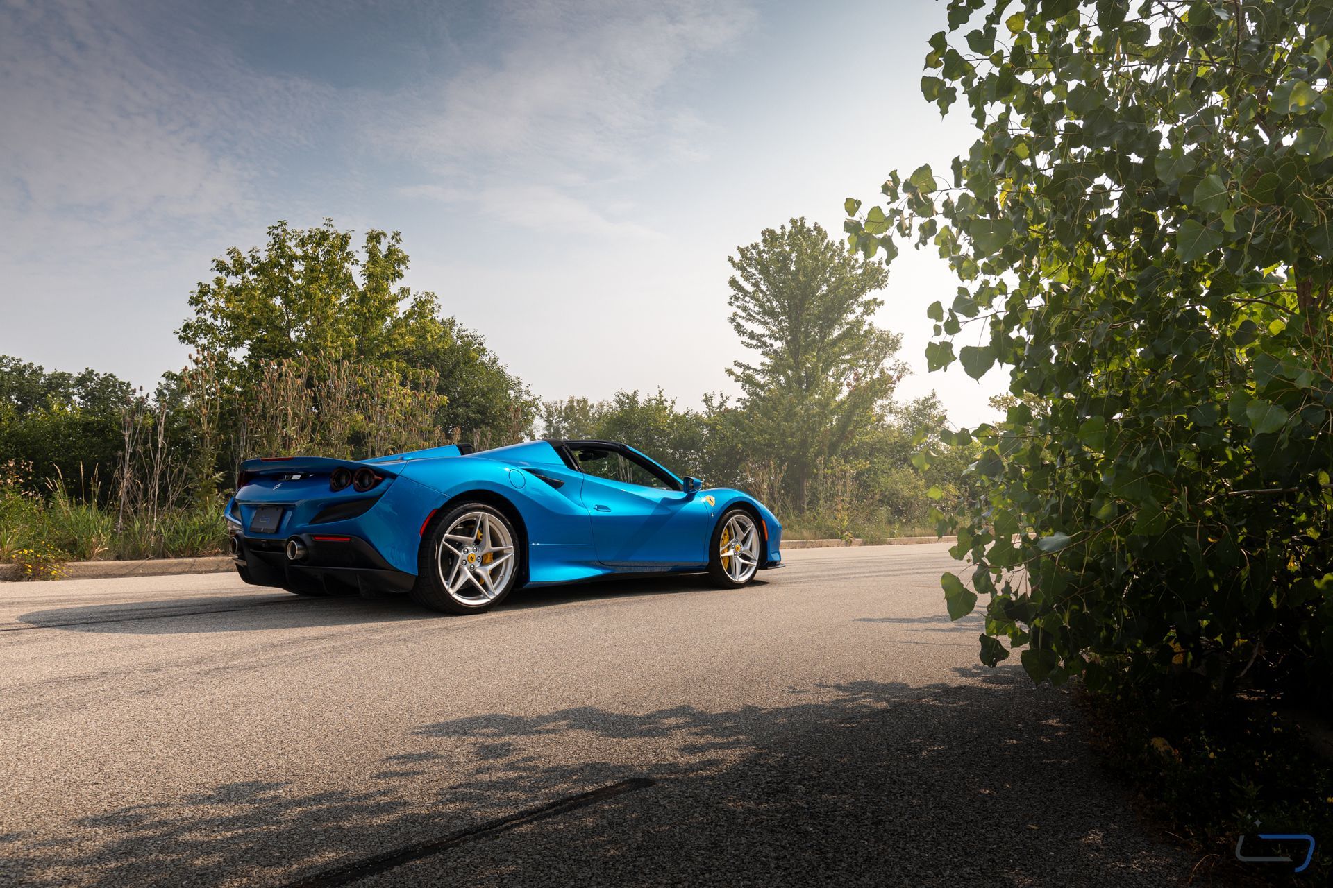 Blue sports car on a sunlit road, framed by trees and bushes.