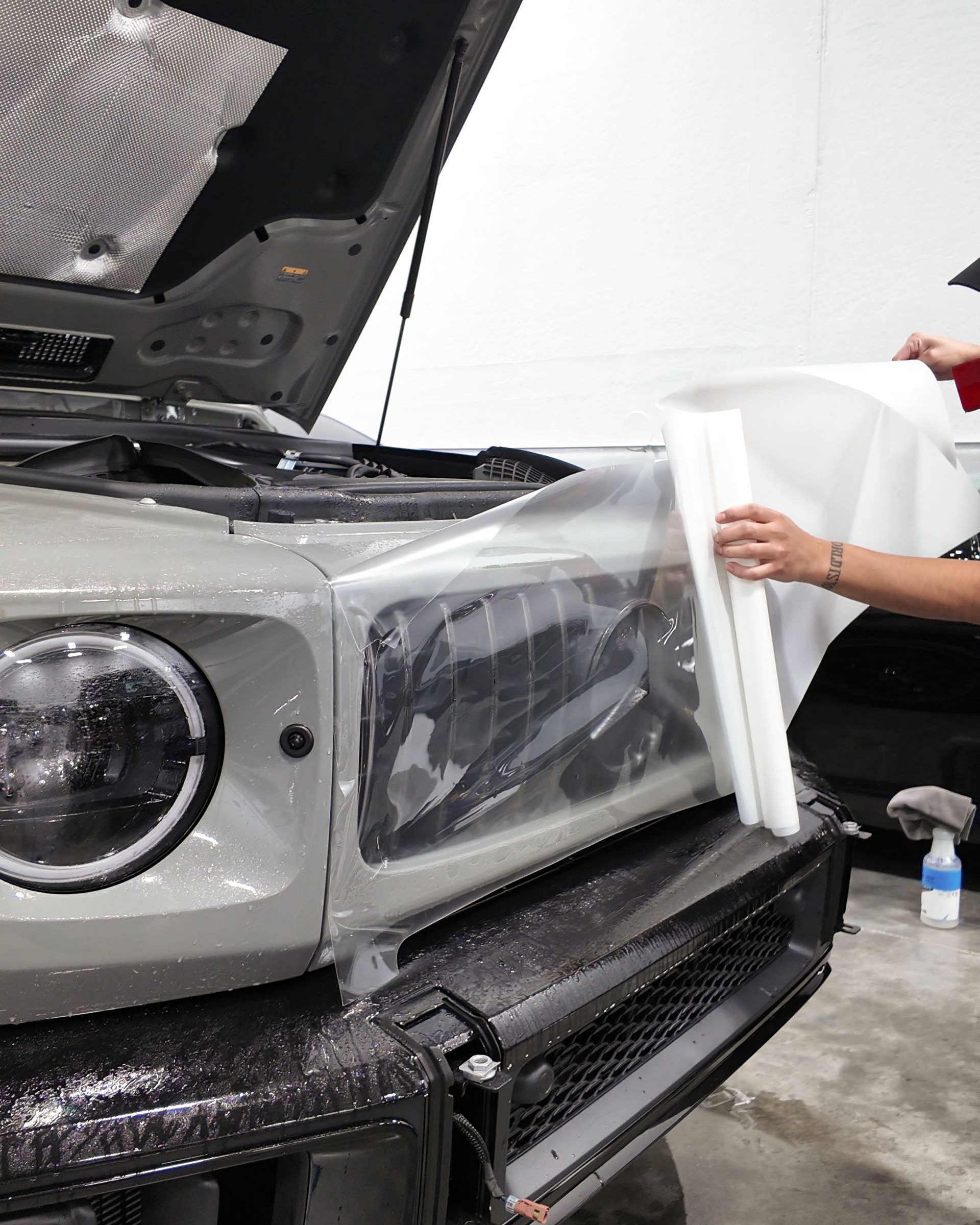 Person applying protective film to the front of a silver car in a workshop.