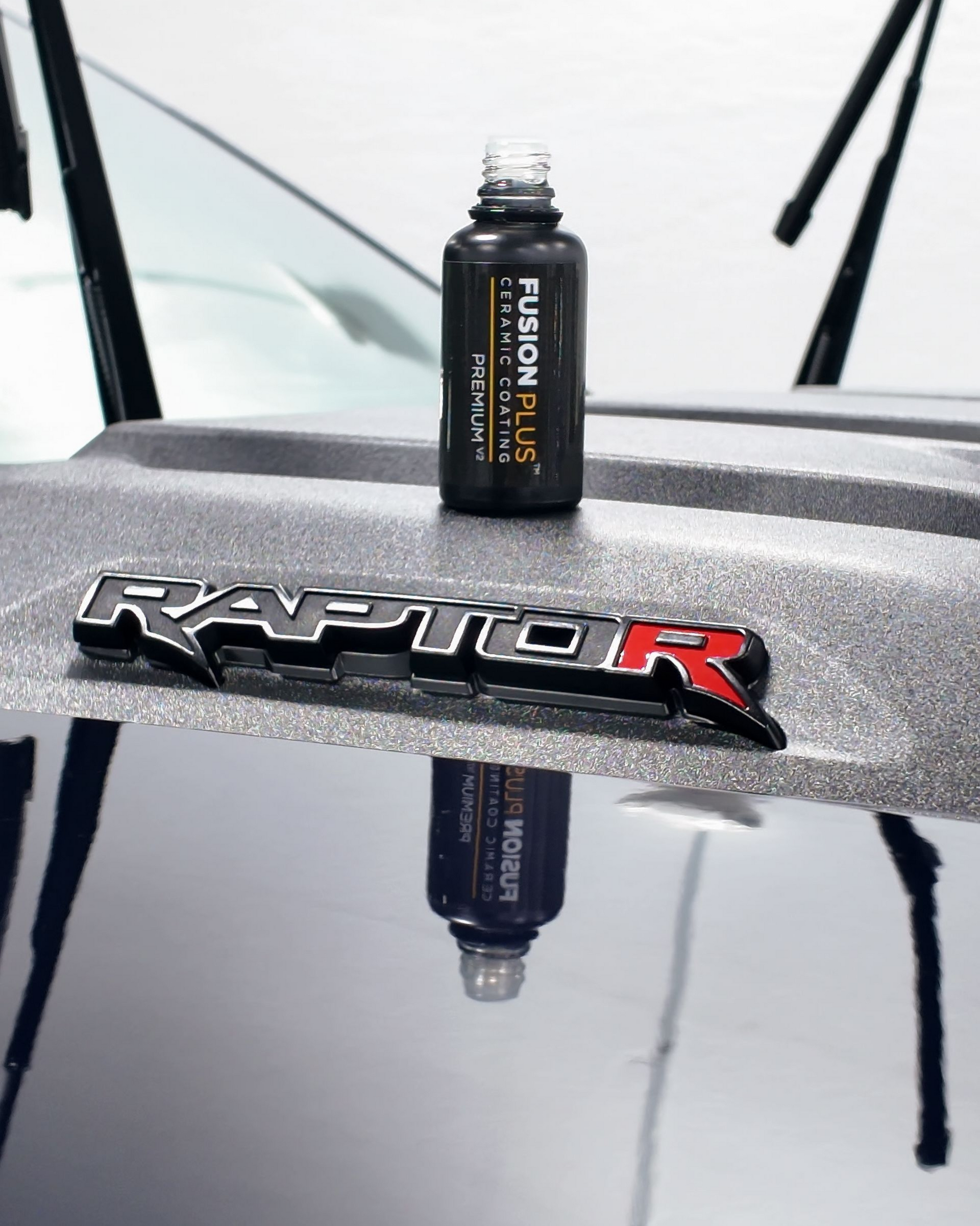 Black bottle on a gray Ford Raptor hood with a reflected logo and windshield wipers in the background