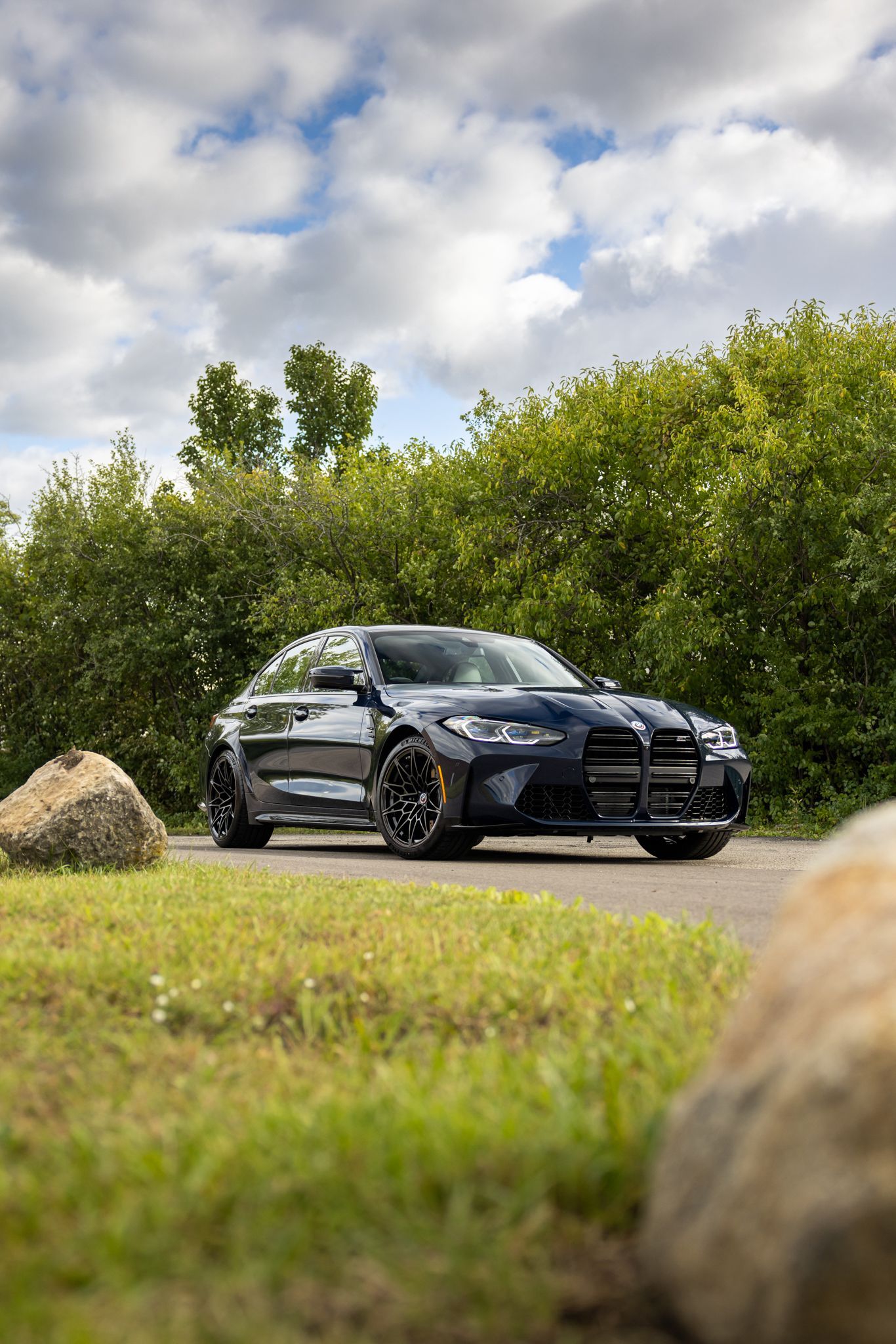 Black luxury sedan parked on a grassy path beside bushes and rocks under a cloudy sky