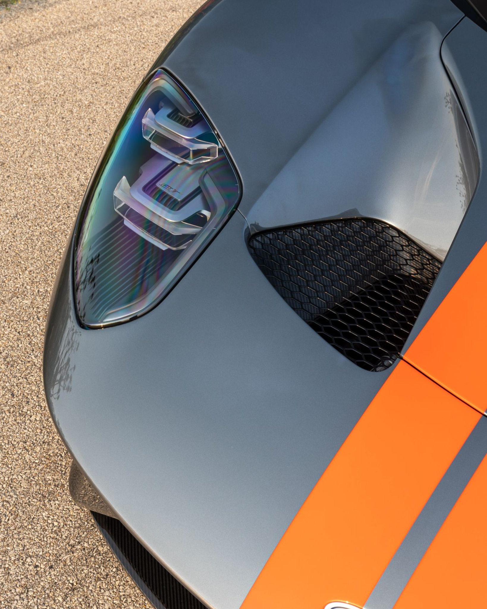 Close-up of a gray sports car with orange racing stripes and a reflective headlight on pavement